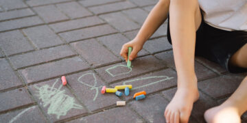 A little preschool boy draws with colorful chalks on the ground. Positive happy toddler child drawing and creating pictures. Creative outdoors activity in summer.